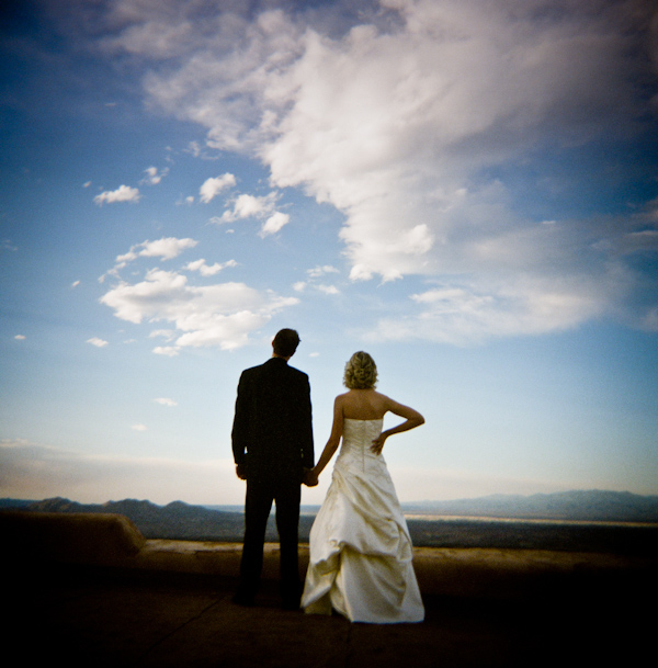photo of the back view of the happy couple holding hands as they look onto a beautiful open landscape and cloud filled sky - bride is wearing a champagne ball gown style dress -  photo by New Mexico based wedding photographers Twin Lens